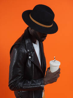 Stylish serious African American male in black leather jacket and hat standing with cup of soda in hand against orange background studio - ADSF46643