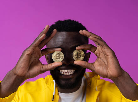 Cheerful young African American male with curly hair smiling while standing and holding bitcoins in fingers in front of eyes against purple background - ADSF46644