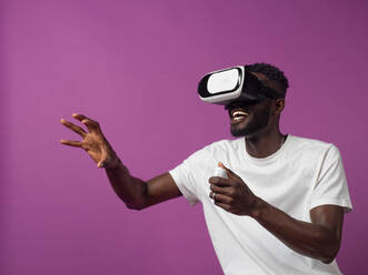 Aufgeregter junger afroamerikanischer Mann in Freizeitkleidung und mit Virtual-Reality-Headset, während er mit halb gefalteten Fingern der erhobenen Hand einen Controller benutzt und ein interessantes Video vor lila Hintergrund betrachtet - ADSF46645