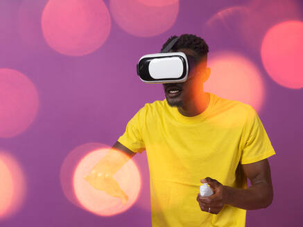 Excited young African American male in casual clothes and virtual reality headset while standing with half folded fingers of hand using controller and watching interesting movie against purple background - ADSF46646