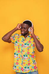 Positive African American male in colorful casual outfit smiling and listening to music through headphones with eyes closed - ADSF46650