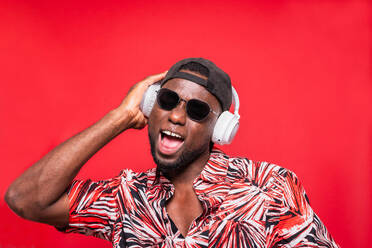 Portrait of man in sunglasses with cap worn backward and listening to music while in open mouth surprised pose with hand son the earphones over red background - ADSF46652