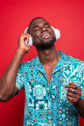 Cheerful African American male in trendy clothes listening audio record music with wireless headset while enjoying songs against red background with eyes closed - ADSF46654