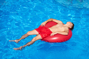 From above full body of positive preteen boy in red shorts lying on red floater over clear blue water swimming pool with eyes closed - ADSF46656
