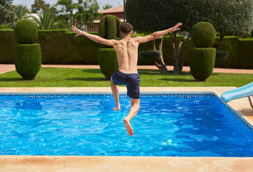 Back view of unrecognizable male in shorts jumping into blue wavy clear swimming pool water while enjoying summer vacation in daylight - ADSF46658