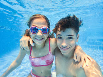 Under water view of happy boy and girl in swimwear and glasses smiling and looking at camera while swimming in blue pool water - ADSF46659