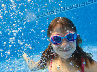 Portrait of confident girl in goggles and swimwear looking at camera while swimming inside water on sunny day - ADSF46660