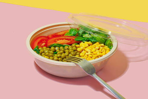 High angle of salad bowl with slices of tomato, spinach leaves, corn kernels and peas placed on pink surface with fork against yellow wall - ADSF46664