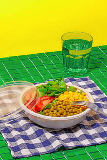 High angle of salad bowl with slices of tomato, spinach leaves, corn kernels and peas placed on napkin on green surface with fork near glass of water against yellow wall - ADSF46665
