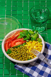 High angle of salad bowl with slices of tomato, spinach leaves, corn kernels and peas placed on napkin on green surface with fork near glass of water - ADSF46666