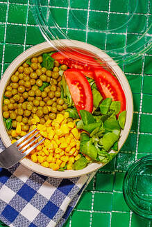Top view of salad bowl with slices of tomato, spinach leaves, corn kernels and peas placed on napkin on green surface with fork near glass of water - ADSF46667