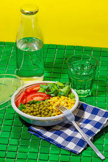 High angle of salad bowl with slices of tomato, spinach leaves, corn kernels and peas placed on napkin on green surface with fork near glass and bottle of water against yellow wall - ADSF46668