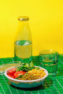 Salad bowl with slices of tomato, spinach leaves, corn kernels and peas placed on green surface near bottle and glass of water against yellow wall - ADSF46669