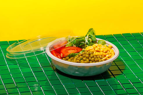 Salad bowl with slices of tomato, spinach leaves, corn kernels and peas placed on green surface against yellow wall - ADSF46670