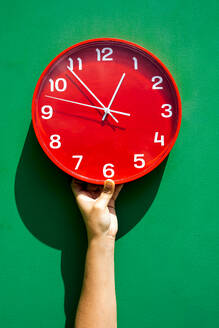 Die Hand einer nicht erkennbaren Person hält eine große runde rote Uhr an einer grünen Wand - ADSF46678