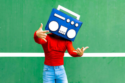 Unrecognizable male in casual clothes covering face with blue boombox showing rock and roll gesture while standing against green background - ADSF46684