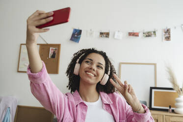 Happy teenage girl showing peace sign and taking selfie through smart phone - ALKF00549