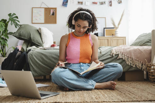 Teenage girl wearing wireless headphones doing homework at home - ALKF00561