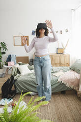 Teenage girl wearing virtual reality simulators gesturing in bedroom at home - ALKF00579