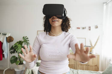 Happy teenage girl wearing virtual reality simulators gesturing in bedroom at home - ALKF00580
