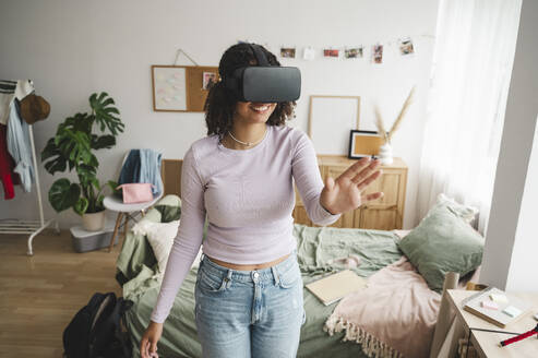 Smiling teenage girl wearing virtual reality simulators gesturing in bedroom at home - ALKF00581