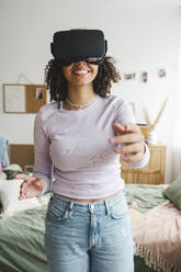 Happy girl wearing virtual reality simulators gesturing in bedroom - ALKF00585