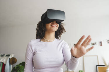 Smiling curly haired girl wearing virtual reality simulators gesturing at home - ALKF00586