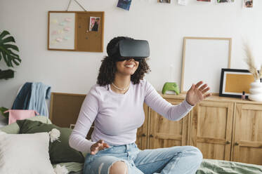 Happy girl wearing virtual reality simulators gesturing on bed at home - ALKF00588