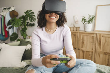 Glückliches Mädchen, das Virtual-Reality-Simulatoren trägt und einen Game-Controller auf dem Bett zu Hause hält - ALKF00592