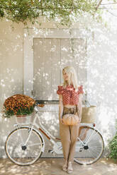 Frau hält Kürbis und steht neben einem Fahrrad mit Herbstblumen - SVKF01605