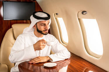 Arabischer Mann mit Kandora im Emirates-Stil fliegt in exklusivem Privatjet - Nahöstlicher Geschäftsmann mit traditioneller Kleidung fliegt in exklusiver Business Class im Flugzeug, Konzepte über Business und Transport - DMDF02851