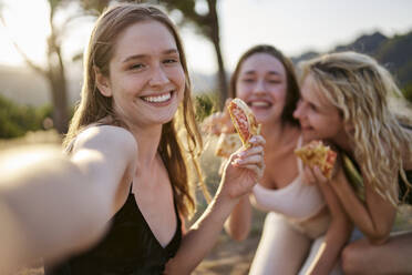 Happy woman having pizza and taking selfie with friends in park - ANNF00362