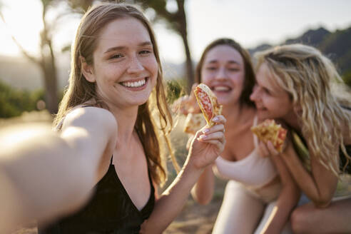 Happy woman having pizza and taking selfie with friends in park - ANNF00362