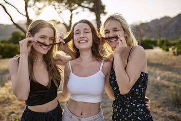 Lächelnde Frauen machen Schnurrbart aus dem Haar eines Freundes im Park - ANNF00370