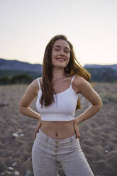 Happy woman with hands on hips standing at beach - ANNF00393