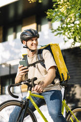 Glücklicher junger Zusteller mit Smartphone auf dem Fahrrad - DAMF01220