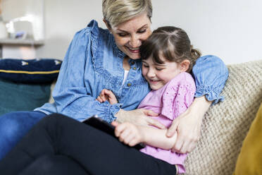 Happy mother embracing daughter holding tablet PC and sitting on sofa - WPEF07563