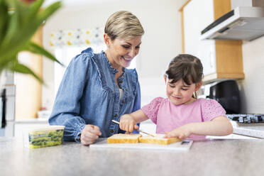 Girl spreading butter on bread by mother standing in kitchen - WPEF07573