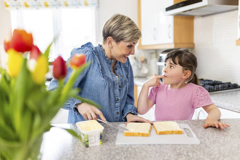Lächelnde Mutter, die Butter auf Brot streicht, während ihre Tochter zu Hause die Finger ableckt - WPEF07574