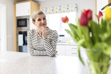 Smiling woman with hand on chin leaning on kitchen counter - WPEF07618