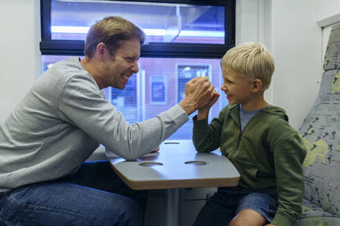 Glücklicher Vater und Sohn beim Armdrücken auf einem Tisch im Zug - NJAF00544