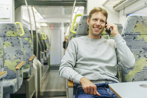 Glücklicher Mann, der im Zug mit seinem Smartphone telefoniert - NJAF00547