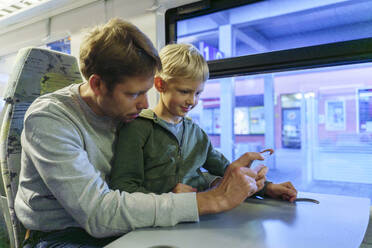 Lächelnder Junge mit Vater, der ein Smartphone in der Nähe des Fensters im Zug benutzt - NJAF00549