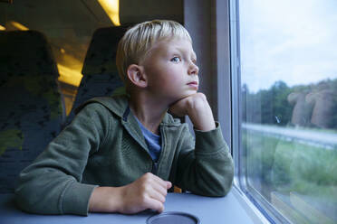 Nachdenklicher Junge mit Hand am Kinn, der durch ein Fenster im Zug schaut - NJAF00554