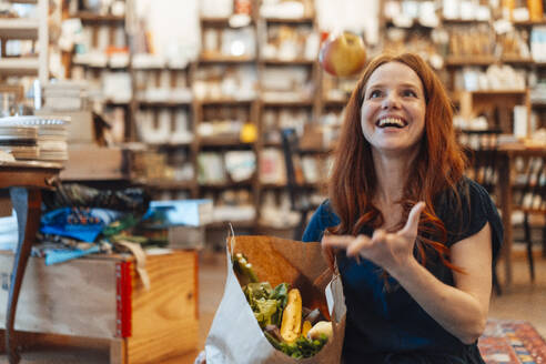 Cheerful woman gesturing and holding bag of groceries at store - KNSF09696