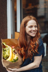 Happy redhead woman standing with groceries bag - KNSF09699