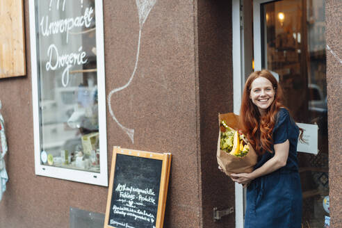 Happy redhead woman standing with groceries bag outside store - KNSF09700