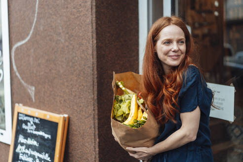 Lächelnde rothaarige Frau mit Einkaufstasche vor dem Laden stehend - KNSF09701