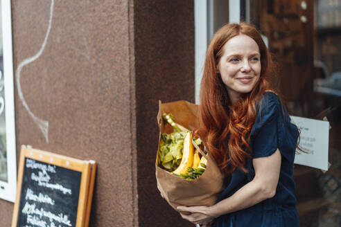 Smiling redhead woman standing with groceries bag outside store - KNSF09701