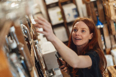 Smiling redhead woman with smart phone reaching for merchandise at store - KNSF09704